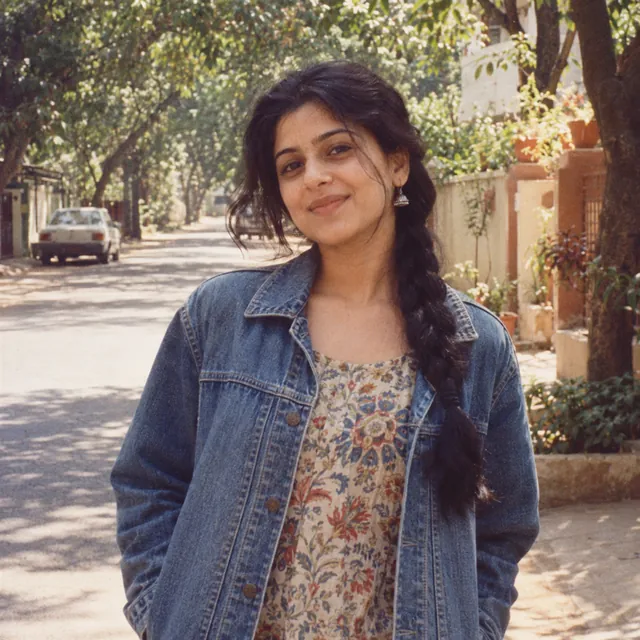1990s film snapshot of a young Indian woman in a denim jacket over a floral top on a leafy residential street