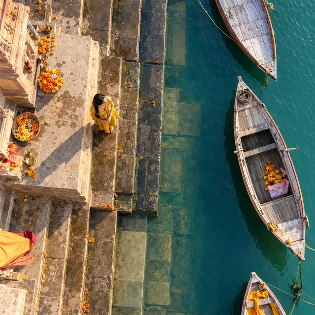 Top-down aerial view of a woman in a mustard kurta on stone river ghat steps beside wooden boats and marigolds