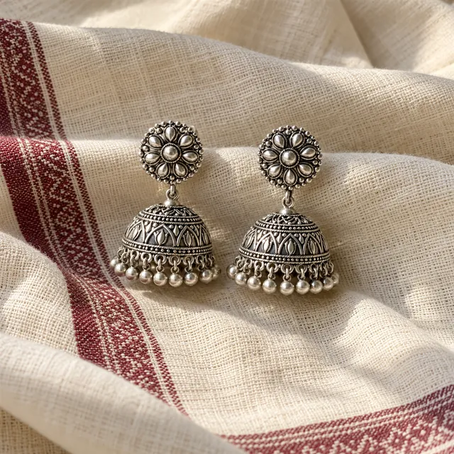Pair of silver jhumka earrings resting on a draped cream handloom cloth with a maroon woven border