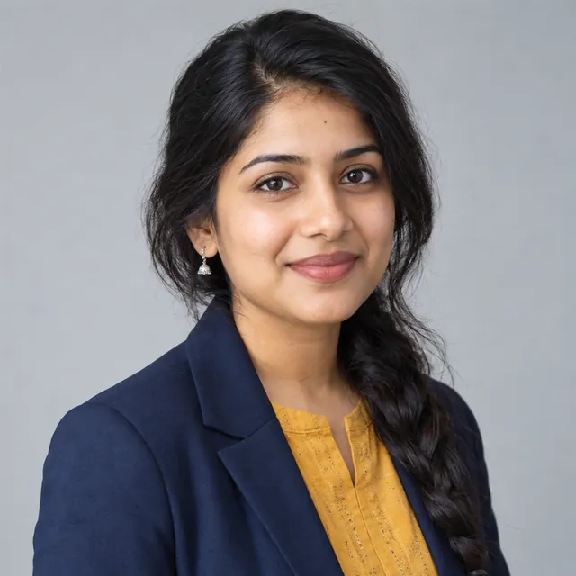 Professional headshot of a young Indian woman in a navy blazer over a mustard kurta, on a plain light-grey background