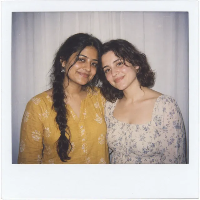 Faded instant-film photo of a woman in a mustard kurta and a female friend smiling in front of a white curtain